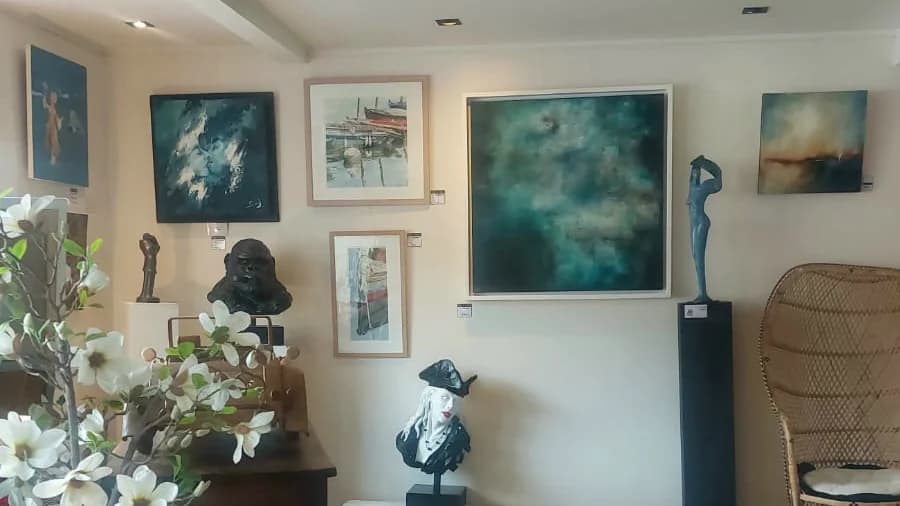 Cimaises de la galerie sous les poutres anciennes : peintures encadrées, sculptures sur socles et fauteuil en rotin